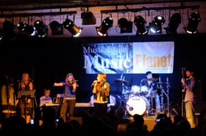 Gitarre, Geige, Keyboard, Schlagzeug und Sängerin auf einer Bühne